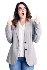 Beautiful young brunette woman wearing business clothes and glasses amazed and surprised looking up and pointing with fingers and raised arms.