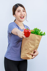 Asian young beautiful housewife in casual wear standing smiling showing and holding brown paper shopping bag of fresh raw organic vegetables from market on white background.