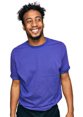 Young african american man with beard wearing casual purple t shirt looking away to side with smile...