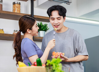 Asian young romantic couple beautiful wife standing smiling using fork feeding sliced mixed fruits from glass bowl to handsome husband behind kitchen counter full of fresh raw organic vegetables