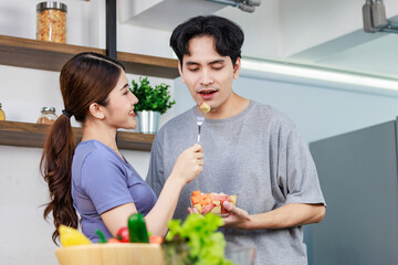 Asian young romantic couple beautiful wife standing smiling using fork feeding sliced mixed fruits from glass bowl to handsome husband behind kitchen counter full of fresh raw organic vegetables
