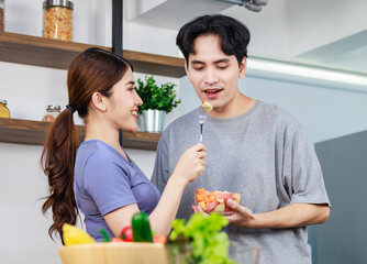 Asian young romantic couple beautiful wife standing smiling using fork feeding sliced mixed fruits from glass bowl to handsome husband behind kitchen counter full of fresh raw organic vegetables