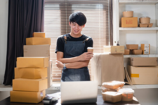 Young Asian man enjoying a small business standing next to a table with notebooks and parcels for delivery to customers crossing his arms and holding cup.