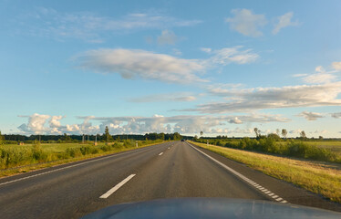 Fototapeta premium Highway (new asphalt road) through the agricultural field and forest at sunset. Dramatic sky after the thunderstorm. Summer, early autumn. Vacations, adventure, road trip, remote places. View from car