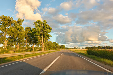 Highway (new asphalt road) through the agricultural field and forest at sunset. Dramatic sky after the thunderstorm. Summer, early autumn. Vacations, adventure, road trip, remote places. View from car