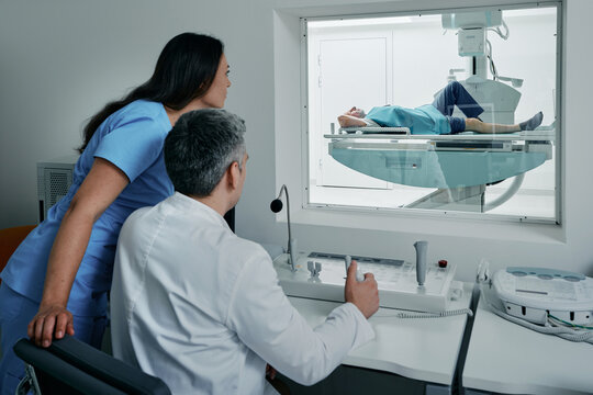 Radiologist with nurse control X-ray process from their office behind protective window, for male patient lying in x-ray machine. X-ray examination