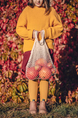 Caucasian teen girl wearing warm knitted clothes staying in autumn park in sunny day and holding in her hands white eco bag with orange pumkins and red apples. Eco lifestyle and healthy eating