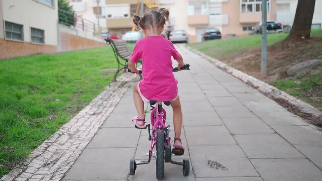 Cute little smiling girl riding bike bicycle in city on parking sunny summer day. Active family leisure with kids. Little caucasian girl 5 years old have fun with pink child bicycle