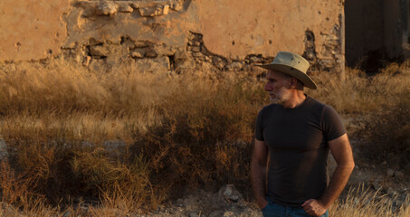 Portrait of adult man in cowboy hat and jeans against abandoned building during sunset