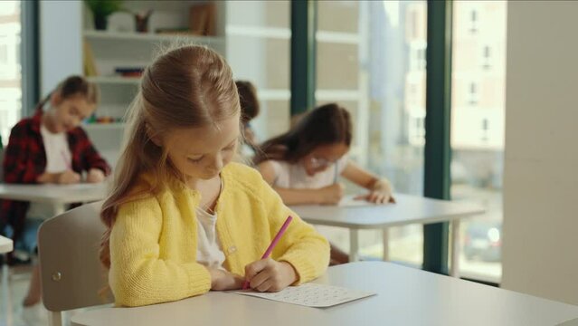 Beautiful long-haired blonde pupil sits at a first desk in the classroom and fills out tests thinks about answers, and the pupils doing test on the background. Back to school.