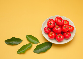 Top view of ripe red acerola cherries on a white dish and green leaves over a yellow background