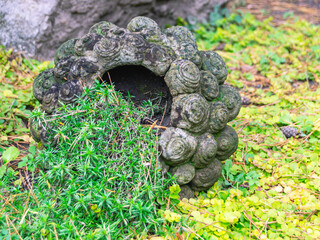 An ancient vase made of gray stone lies on the grass. Landscape design. A green plant grows from an old vase.