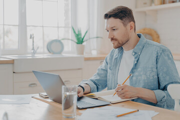 Concentrated young businessman writing notes on agenda and working remotely on laptop