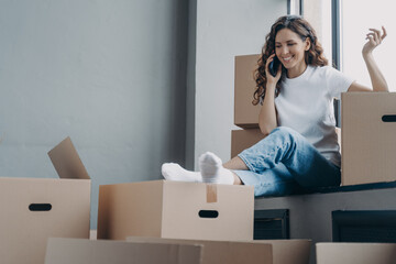 Happy hispanic woman sitting on windowsill of new house. Girl unpacking boxes and talking on phone.