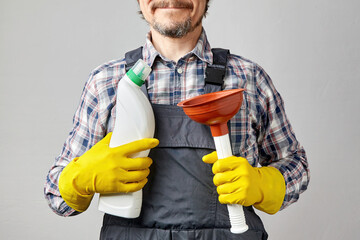 A caucasian man with a mustache and beard in overalls holds a plunger and a bottle of pipe cleaner...