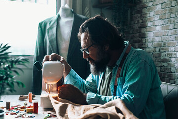 One man in home workplace use sewing machine to work on vintage garment alone. Male people working as a tailor indoor. Alternative business store concept lifestyle. Young adult in garment production