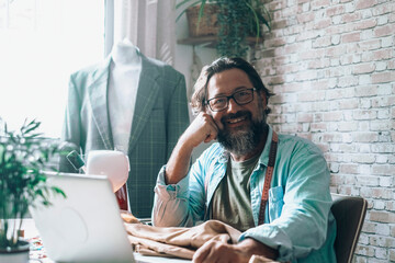 Portrait of a tailor sitting at the desk with laptop and sewing machine. Modern businessman tailor...