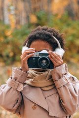 Cute African-American girl in fur headphones takes pictures with a camera in an autumn park.Diversity,autumn concept.