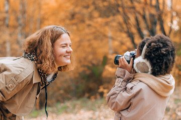 African-American daughter takes pictures of her caucasian mother on camera in an autumn park.Diversity,autumn concept.Side view,selective focus.