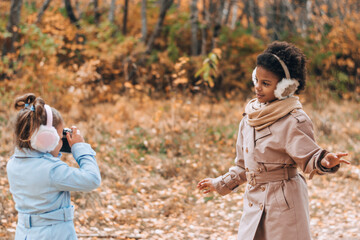 Caucasian and African-American girls take pictures of each other on camera in the autumn park.Technology,diverse people,autumnal concept.