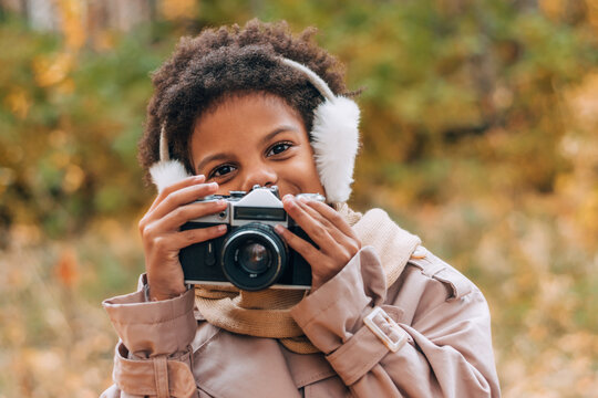 Cute happy African-American girl in fur headphones takes pictures with a camera in an autumn park.Diversity,autumn concept.