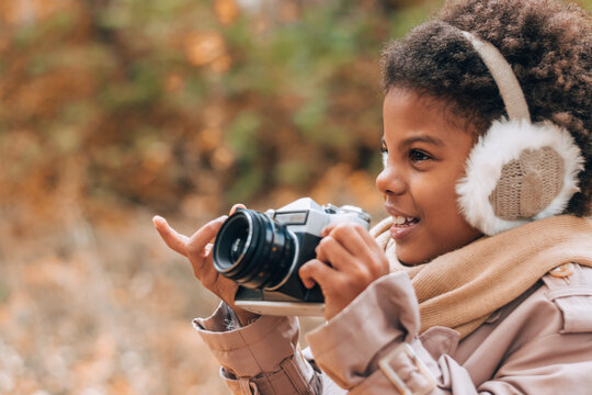 Cute happy African-American girl in fur headphones takes pictures with a camera in an autumn park.Diversity,autumn concept.Copy space.