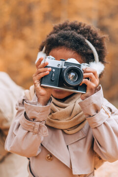 Cute African-American girl in fur headphones takes pictures with a camera in an autumn park.Diversity,autumn concept.