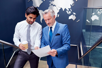 Male Colleagues Meeting On Stairs Of Office Discussing Document With World Map In Background