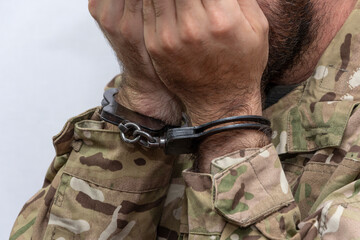 A soldier in military uniform and handcuffs covers his face with his hands on a light background.Concept: war criminal, army court and tribunal, sentencing for a prisoner.