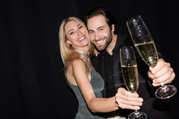 Blonde woman in dress hugging boyfriend and holding glass of champagne isolated on black.