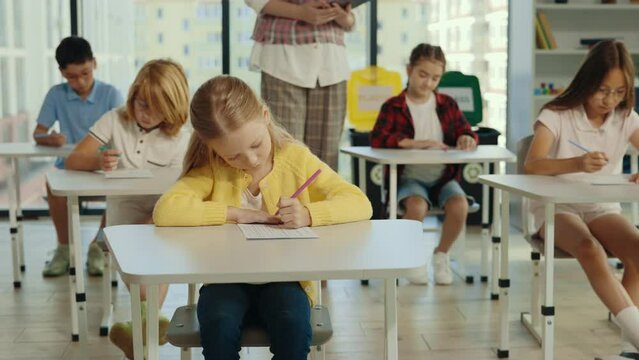 Back to school. Beautiful pupil sits at a first desk in the classroom and fills out tests, and the teacher in the background help students with answers. People and educational concept.
