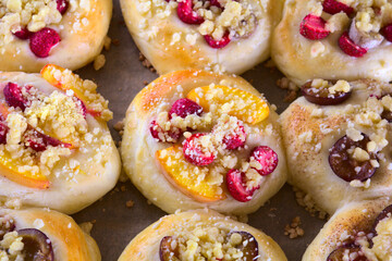 Closeup of freshly baked homemade sweet rolls with various seasonal fruits (strawberries, nectarines, plums, bananas) and golden crumble. The rolls are placed on a baking tray lined with paper.