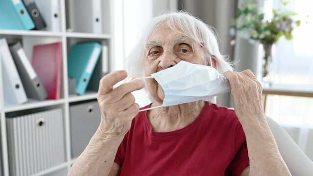 Senior woman takes off medicine mask at home in pandemic time. Elderly woman with protection from coronavirus covud flu