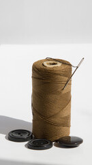 Brown threads on a spool along with a needle and buttons next to it on a white background