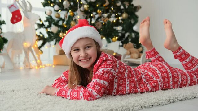 Happy child girl lying on floor near to Christmas tree wearing red Santa hat and smiling. Beautiful female kid enjoying New Year and Xmas time