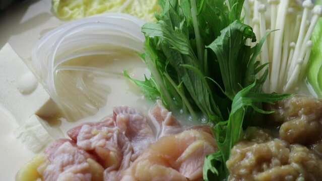 鶏もも肉、鶏つくね、水菜、豆腐を白濁した鶏白湯スープで煮込む鍋料理。土鍋で加熱される具材を捉えた接写・クローズアップ映像。飲食店メニュー、レシピ解説、冬の料理、和食の広告や映像素材に。 | Close-up of chicken and vegetable hot pot in creamy white soup.