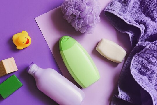 Green shampoo bottle, purple shower gel packaging, towel, sponge and yellow rubber duck. Baby care cosmetic products flat lay photography