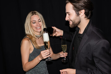 Smiling man in jacket pouring champagne near blonde girlfriend during party isolated on black.