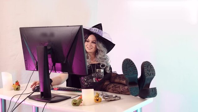 Beautiful witch sits at the computer with halloween decorations, working on halloween typing on the keyboard and surfs the web, drinks a little and smiles, is surprised by an employee