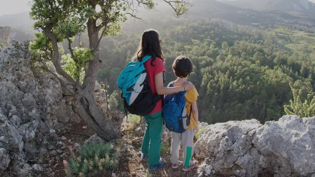 The boy and his mother are standing on the top of the mountain, A woman is traveling with child, Boy with his mother looking at the mountains, Travel with backpacks, Hike and climb with kids