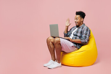 Full body young man of African American ethnicity wear blue shirt sit in bag chair use work on laptop pc computer waving hand isolated on plain pastel light pink background. People lifestyle concept.