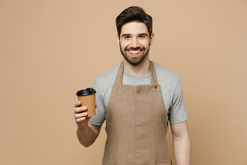 Young cheerful happy man barista barman employee wear brown apron work in shop hold craft paper brown cup coffee to go isolated on plain pastel light beige background. Small business startup concept.