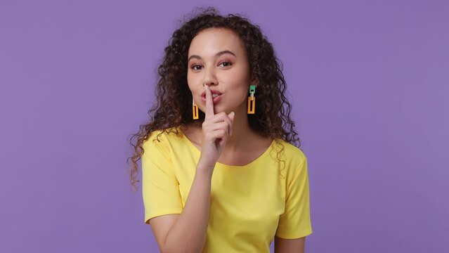 Young secret woman of African American ethnicity 20s wear yellow t-shirt look aside say hush be quiet with finger on lips shhh gesture isolated on plain pastel light purple background studio portrait