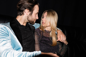 Stylish man hugging blonde girlfriend on leather couch during party isolated on black.