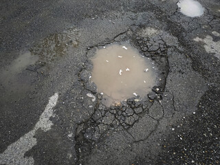 The paved road surface is damaged. Broken asphalt. Puddle on cracked road.