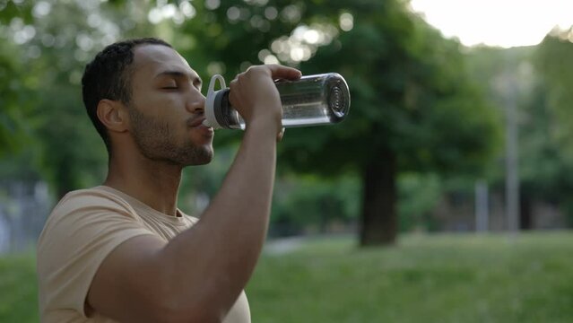Side view of african american man is drinking water from the sport bottle. Male runner standing in the city park after jogging. Sport and healthy lifestyle concept.