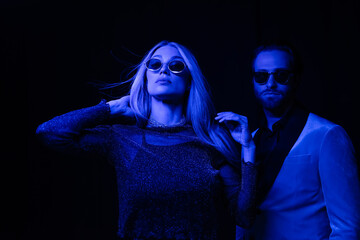 Trendy blonde woman standing near boyfriend in sunglasses isolated on black with blue lighting.
