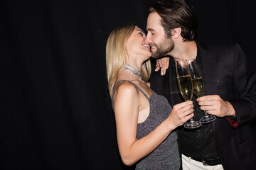 Side view of smiling stylish couple kissing while holding champagne isolated on black.