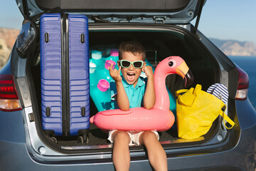 cheerful boy in sunglasses is sitting in the trunk with a suitcase, an inflatable circle.