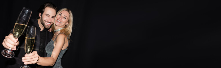 Cheerful stylish couple hugging and holding glasses of champagne isolated on black, banner.